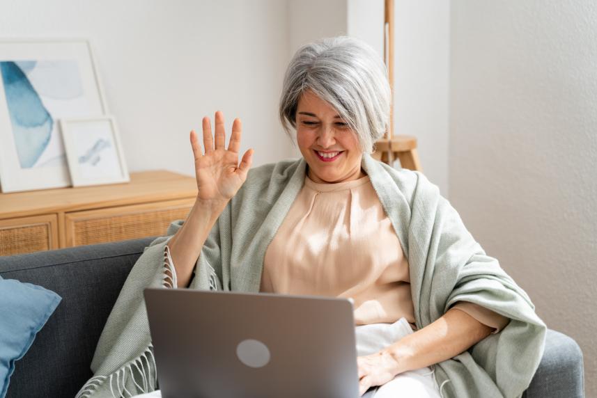 An older woman with gray hair waves at her laptop camera during a telehealth visit.