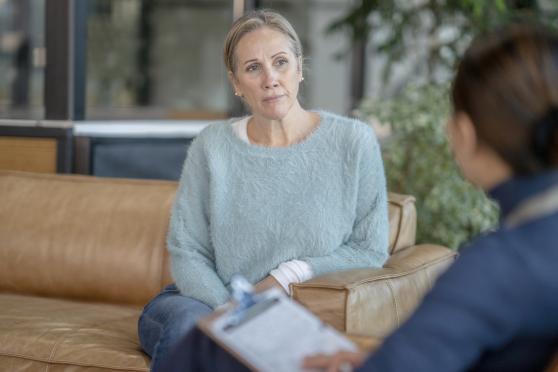 A woman in a light blue sweater sits on a sofa during an appointment with a mental health provider.